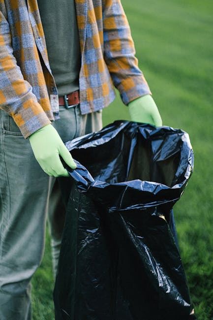 A person dressed in casual outdoor clothing, including a plaid shirt and green gloves, is holding open a large black rubbish bag lined with a grey interior, prepared for waste disposal. The individual