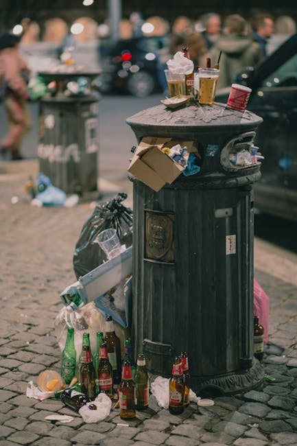 A narrow alleyway flanked by tall brick and concrete buildings on either side, with graffiti on the right wall. In the foreground, a large, grey, weathered rollable waste container with a lid stands o